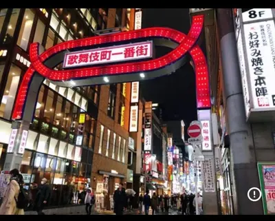a city street with a large red sign with people at \u65b0\u5bbf\u99c52\u5206 jr\u65b0\u5927\u4e45\u4fdd\u99c5\u5f92\u6b698\u5206 \u6b4c\u821e\u4f0e\u753a\u5f92\u6b6910\u5206 in Tokyo